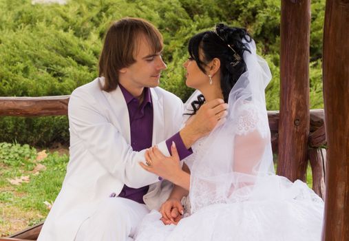 Couple in love bride and groom posing sitting on wooden bench in gazebo in their wedding day in summer.