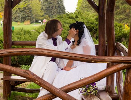 Couple in love bride and groom posing sitting on wooden bench in gazebo in their wedding day in summer.