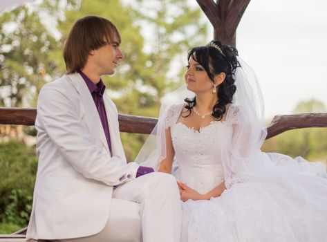 Couple in love bride and groom posing sitting on wooden bench in gazebo in their wedding day in summer.