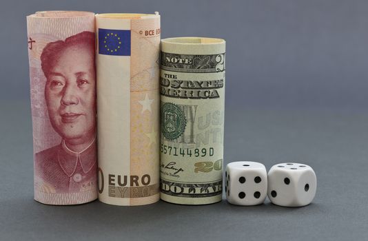 Financial investment risks reflected in dice next to dollar, euro, and yuan money on gray and blue hued background.  Three currencies build concept of global marketplace and  shared gambles. 