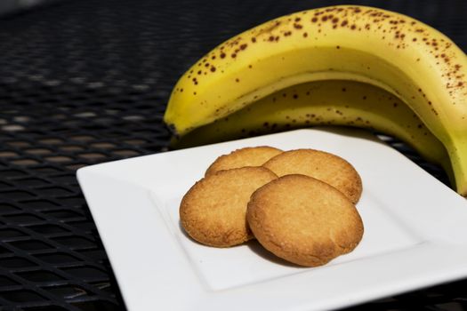 Healthy snack of tasty butter cookies and ripe bananas.  Four cookies on white plate in front of fresh fruit.  Items on outdoor, black metal patio table. 