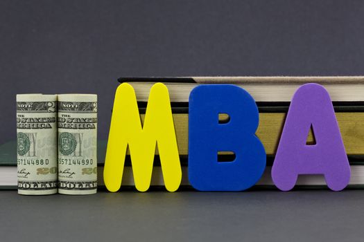 Graduate degree MBA letters with dollar currency in front of books on gray background.  Advance degree is a education business investment in critical skills.  