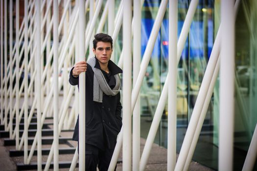 Stylish Young Handsome Man in Black Coat Standing in City Center Street among White Metal Bars of Moden Architecture