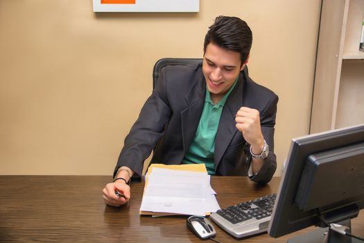 Happy young businessman joyful for success, sitting at desk in his office working with computer