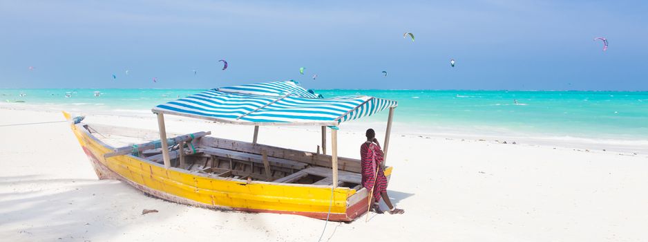 Maasai warrior lounging aroundon traditional colorful wooden boat on picture perfect tropical sandy beach on Zanzibar, Tanzania, East Africa. Kiteboarding spot on Paje beach.