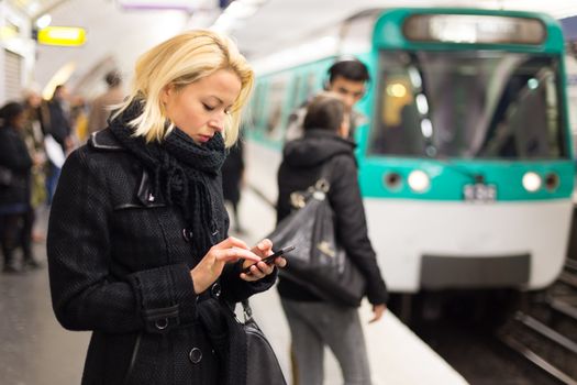 Young woman in winter coat with a cell phone in her hand waiting on the platform of a railway station for train to arrive. Public transport.  