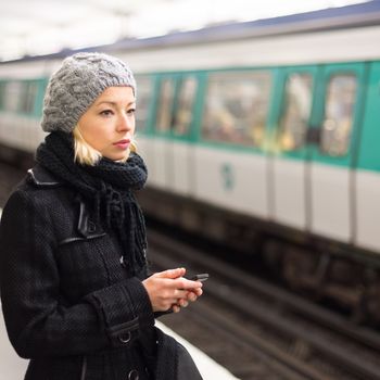 Young woman in winter coat with a cell phone in her hand waiting on the platform of a railway station for train to arrive. Public transport.  