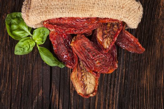 Delicious dried tomatoes with fresh basil leaves in burlap bag on brown wooden vintage textured background. Traditional mediterranean kitchen.