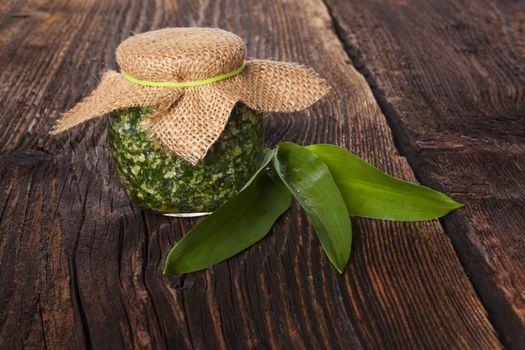 Garlic pesto in glass jar with fresh wild garlic leaves on brown wooden textured background. Culinary healthy eating. 