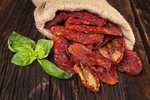 Delicious dried tomatoes with fresh basil leaves in burlap bag on brown wooden vintage textured background. Traditional mediterranean kitchen.