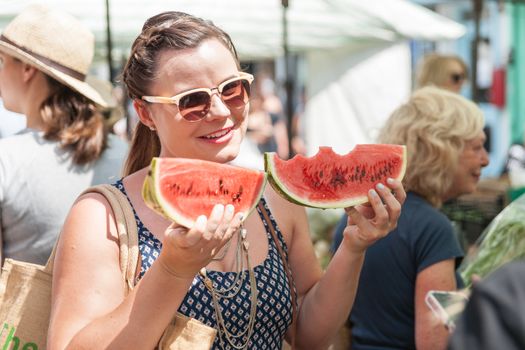 Woman at London Saturday market holds two fresh red watermelon slices, one with three bites.