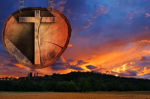 Wooden Christian cross on a section of tree trunk, hanging from a metal chain at the beautiful sunset over the hills with clouds