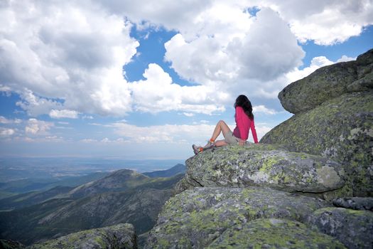 trekking red jacket brown shorts woman sitting back on rock top summit looking at valley at Gredos mountains in Avila Spain Europe
