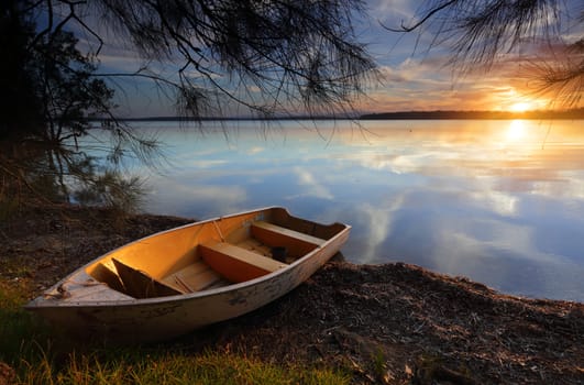 Sublime scene and tranquil waters reflecting like a mirror as the divine sun sets casting its golden orange glow upon the clouds and highlighting the little boat as it rests on the shoreline, waiting for tomorrows dreams and adventures.