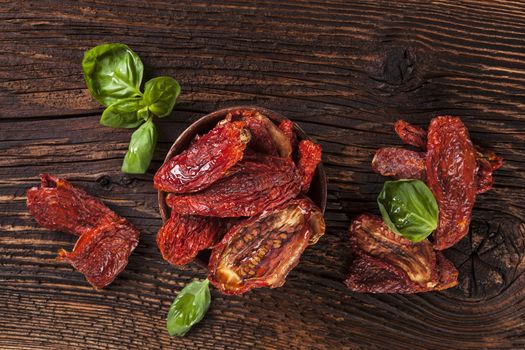 Delicious dried tomatoes with fresh basil herbs on brown wooden vintage textured background, top view. Traditional mediterranean kitchen.