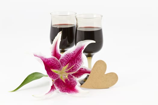 An elegant lily blossom placed with two glasses of red wine and a gold heart on white background; horizontal image with copy space.