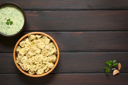 Cooked tortellini stuffed with cheese in wooden bowl with parsley cream sauce, photographed overhead on dark wood with natural light