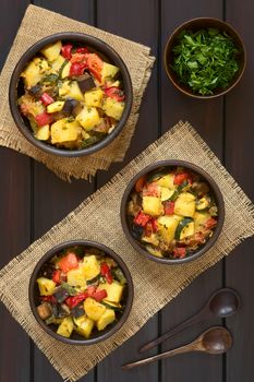 Baked potato, eggplant, zucchini and tomato casserole in rustic bowls, photographed overhead on dark wood with natural light