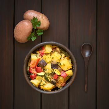 Baked potato, eggplant, zucchini and tomato casserole in rustic bowl, raw potatoes, parsley leaf and wooden spoon on the side, photographed overhead on dark wood with natural light