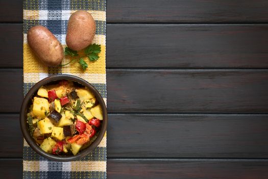Baked potato, eggplant, zucchini and tomato casserole in rustic bowl, with raw potatoes and parsley leaf on kitchen towel, photographed overhead on dark wood with natural light
