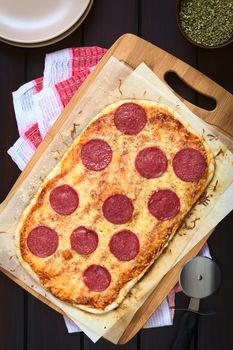 Homemade pepperoni or salami pizza on baking paper on wooden board with plates, dried oregano and pizza cutter on the side, photographed overhead on dark wood with natural light