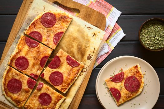 Homemade pepperoni or salami pizza cut in pieces on baking paper on wooden board, photographed overhead on dark wood with natural light