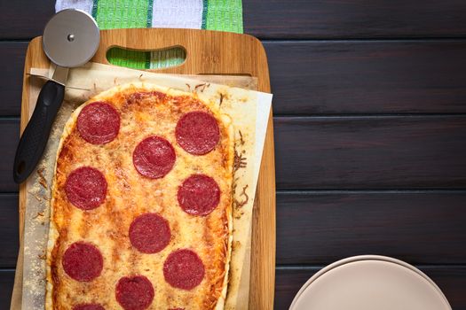 Homemade pepperoni or salami pizza on baking paper with pizza cutter on wooden board, photographed overhead on dark wood with natural light