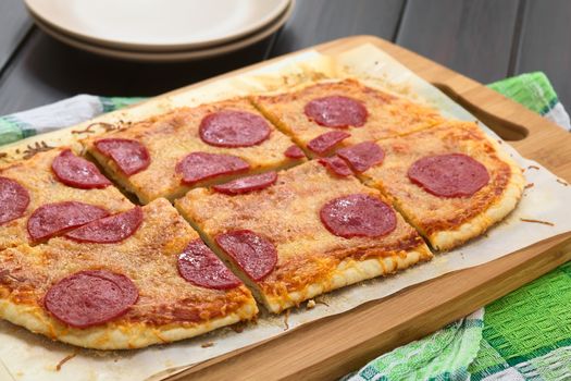 Homemade pepperoni or salami pizza cut in pieces served on baking paper on wooden board, photographed on dark wood with natural light (Selective Focus, Focus one third into the image)