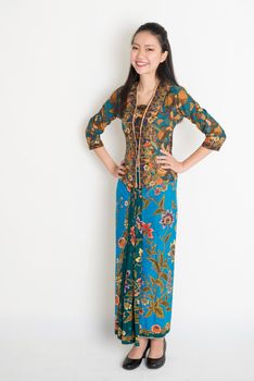 Full length Southeast Asian woman in batik dress standing on plain background.