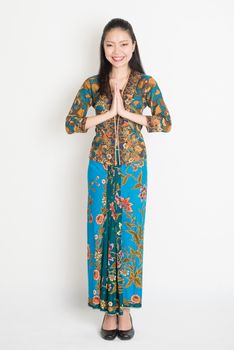 Full length Southeast Asian woman with batik dress in greeting gesture standing on plain background.