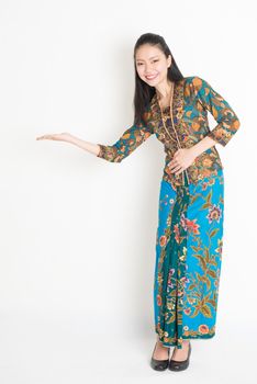 Full length portrait of Southeast Asian girl in batik dress hand holding something standing on plain background.