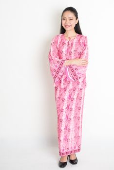 Full body portrait of happy Southeast Asian woman in pink batik dress standing on plain background.