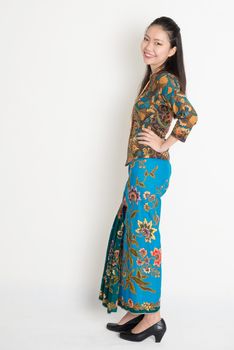 Full length cheerful Southeast Asian female in batik dress standing on plain background.