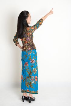 Full body rear view of Southeast Asian woman in batik dress finger pointing away, standing on plain background.