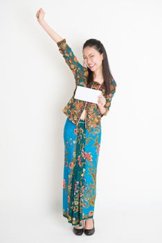 Full length portrait of Southeast Asian female in batik dress hands holding an envelope smiling, standing on plain background.