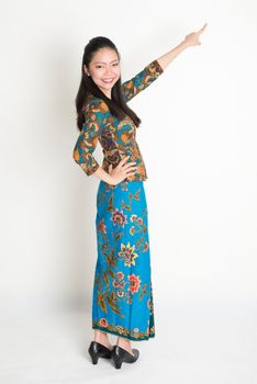 Full body portrait of happy Southeast Asian woman in batik dress finger pointing away, standing on plain background.