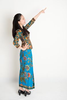 Full body side view of happy Southeast Asian woman in batik dress finger pointing away, standing on plain background.