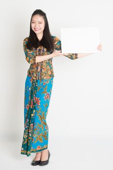 Full body portrait of Southeast Asian girl in batik dress hands holding white blank card, standing on plain background.