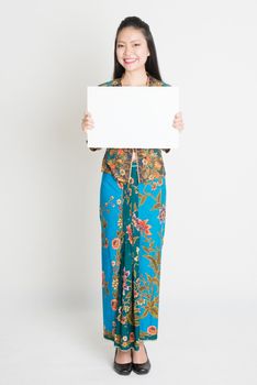 Full body portrait of Southeast Asian girl in batik dress hands holding white blank placard, standing on plain background.