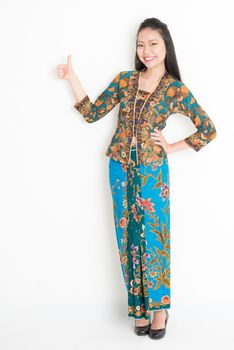 Full body portrait of Southeast Asian woman in batik dress giving thumb up, standing on plain background.
