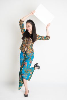 Full body portrait of Southeast Asian girl in batik dress hands holding white blank card jumping around on plain background.