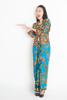 Full body portrait of excited Southeast Asian girl in batik dress hand holding something, standing on plain background.