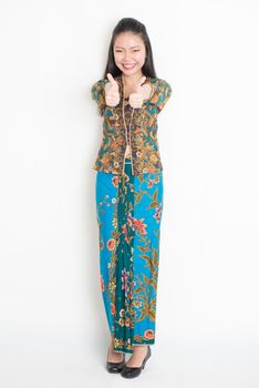 Full length portrait of Southeast Asian woman in batik dress thumbs up standing on plain background.