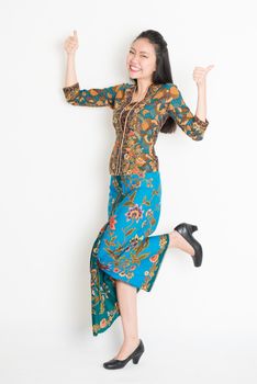 Full length portrait of Southeast Asian woman in batik dress thumbs up, cheering and jumping around on plain background.