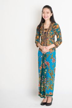 Full length Southeast Asian female in batik dress standing on plain background.