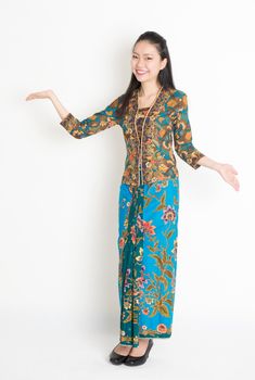 Full length portrait of Southeast Asian woman in batik dress showing welcome gesture standing on plain background.