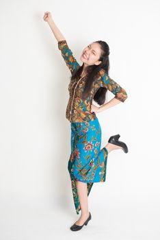 Full length portrait of Southeast Asian female in batik dress arms up, cheering and jumping around on plain background.