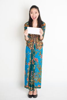 Full length portrait of cheerful Southeast Asian female in batik dress hands holding an envelope standing on plain background.