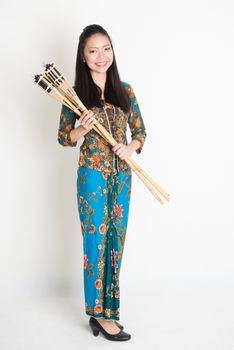 Full body portrait of Southeast Asian girl in batik dress hands holding oil lamp torch standing on plain background.