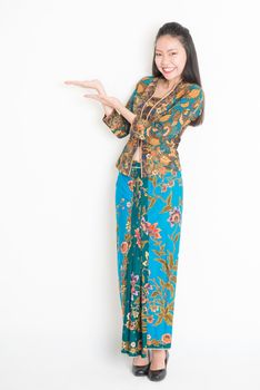 Full body portrait of Southeast Asian female in batik dress hand holding something standing on plain background.